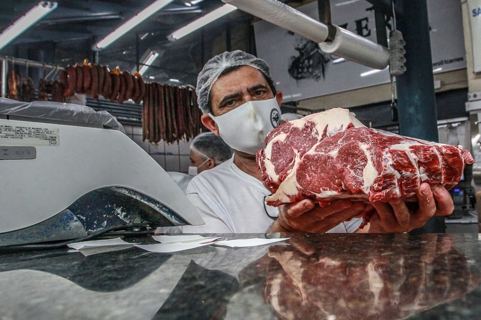 Açougueiro mostra, com as duas mãos, alguns pedaços de carne, apoiando-se no balcão, onde há uma balança