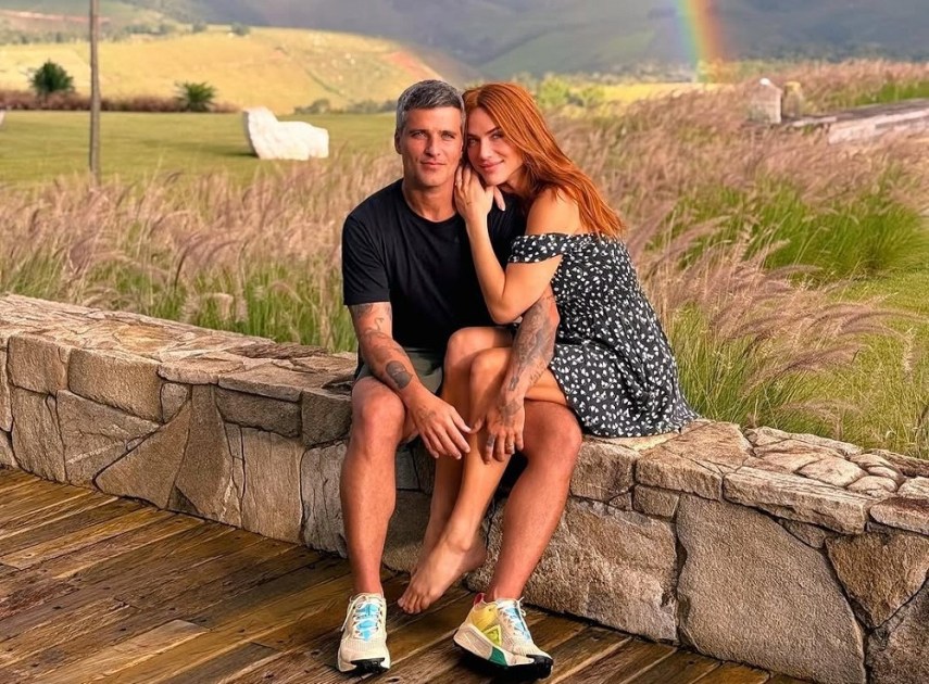 Bruno Gagliasso e Giovanna Ewbabnk sentados em área aberta, com arco-íris ao fundo