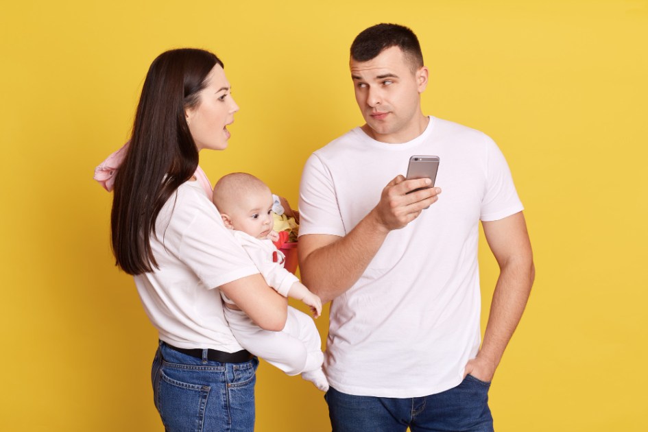 Jovem mãe furiosa segurando o bebê nas mãos e olhando para o marido usando o telefone