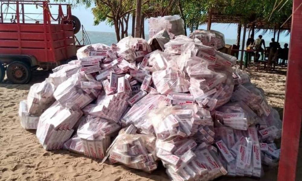 A imagem mostra uma grande pilha de sacos empilhados em uma praia, com areia ao redor. Os sacos são de cor clara e estão amarrados com fitas vermelhas. Ao fundo, há uma estrutura de palha que parece ser um quiosque, e algumas árvores verdes. Um veículo vermelho está visível à esquerda da imagem. Parte de carga de cigarros contrabandeados que estava em embarcação que encalhou em Cajueiro da Praia, litoral do Piauí