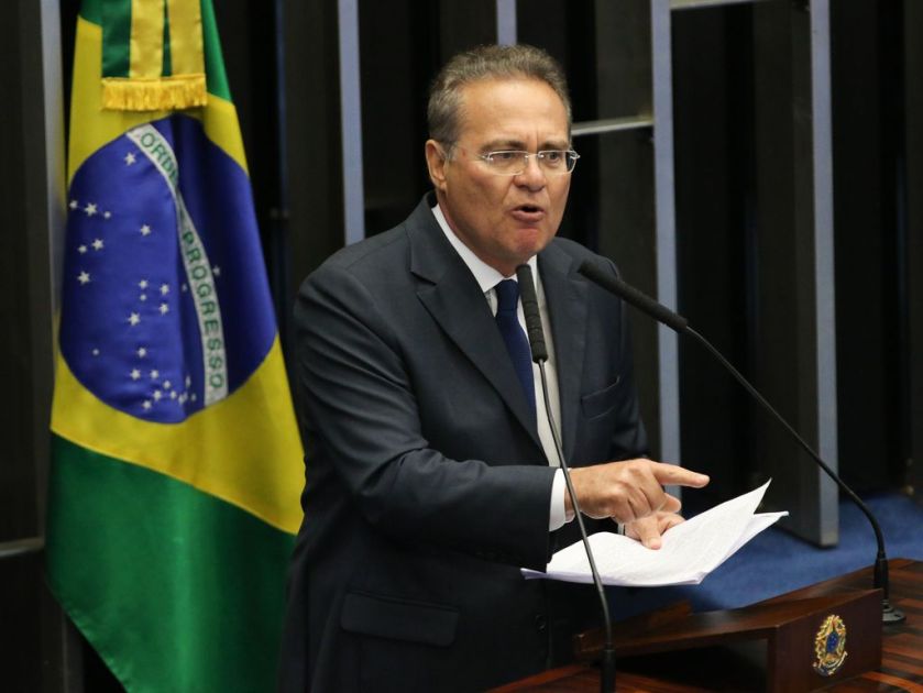 Senador Renan Calheiros discursando em tribuna, com um papel na mão e em frente a um microfone. Ele tem o dedo levantado e está falando. É um homem branco de óculos, com cabelos grisalhos e usa terno preto.