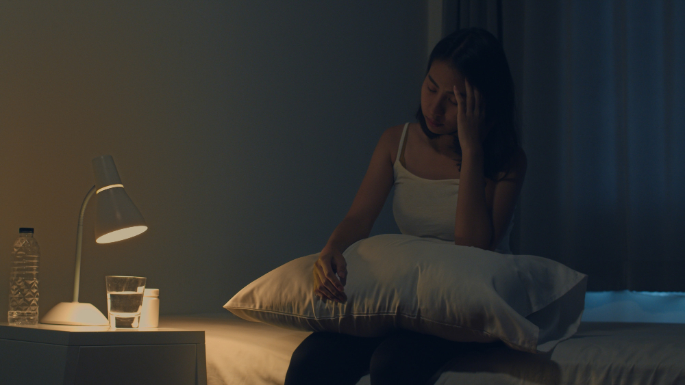 A jovem mulher asiática bonita que senta-se na cama toma o comprimido para dormir ou o remédio da noite no quarto. A mulher indiana doente e insalubre sofre de insônia ou dor de cabeça, a menina deprimida tem remédios antidepressivos.