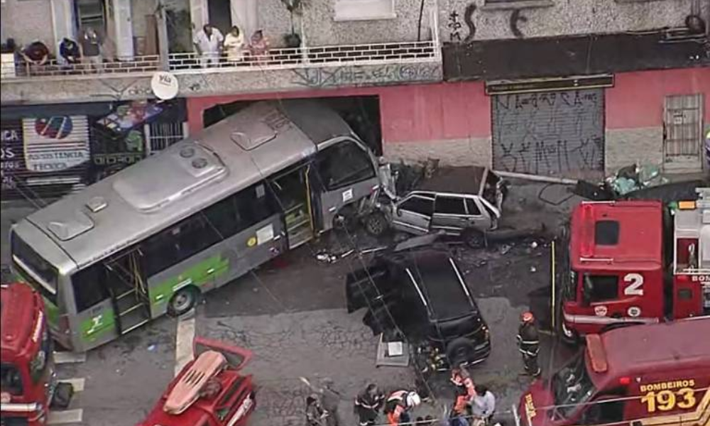 Ônibus colidido e tombado parcialmente na calçada, com dois carros amassados próximos. Equipes de bombeiros e veículos de emergência no local atendem a ocorrência. Pessoas observam da calçada e sacada de prédio próximo. Após colisão, micro-ônibus invadiu um comércio, na zona norte de São Paulo - Reprodução/TV Globo