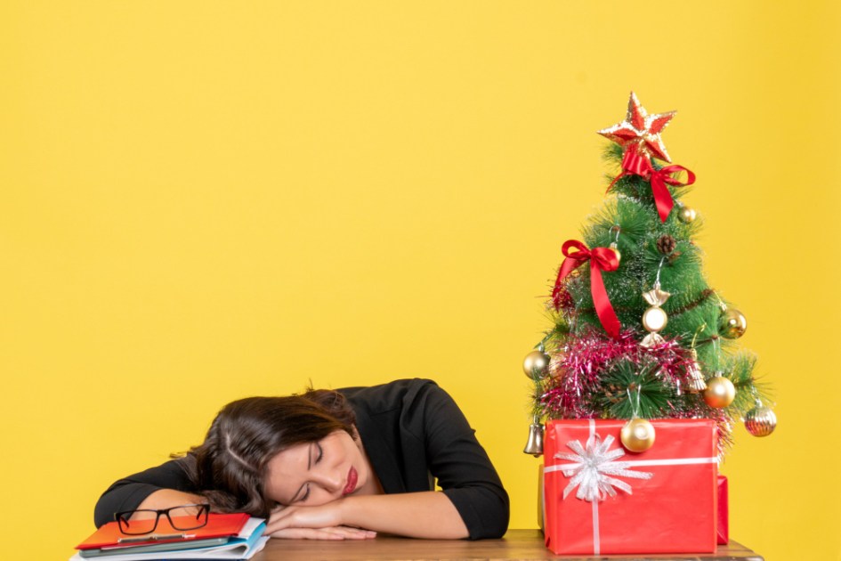 Mulher jovem dormindo sentada em uma mesa perto da árvore de Natal decorada no escritório em amarelo