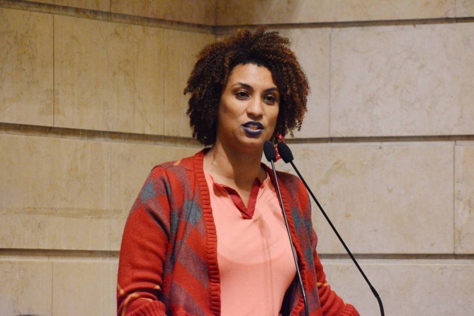 marielle franco falando em um microfone. Mulher negra de cabelos crespos castanhos e batom preto, com camisa rosa e casaco vermelho e azul